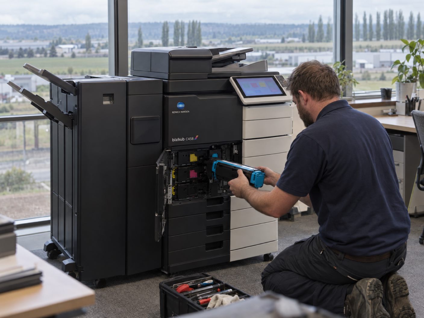 A technician kneeling at the open front of a Konica Minolta bizhub C458 copier, fitting a cyan toner cartridge, in an office above a flat valley of warehouse units and a line of poplars.