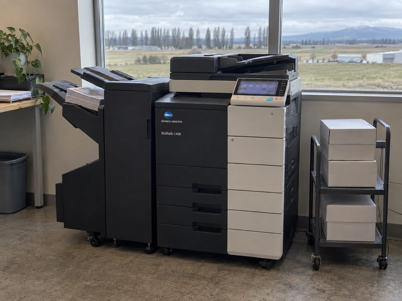 A Konica Minolta bizhub C458 copier standing ready with printed colour pages in its output tray, in an office above a flat valley of warehouse units and a line of poplars.