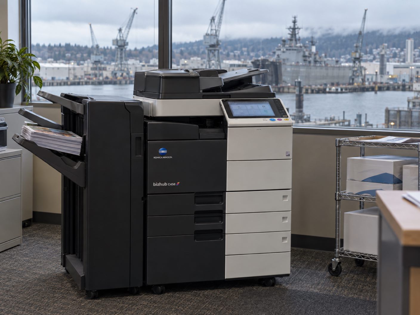 A Konica Minolta bizhub C458 copier standing ready with printed colour pages in its output tray, in an office over a working naval waterfront of shipyard cranes and a grey vessel.