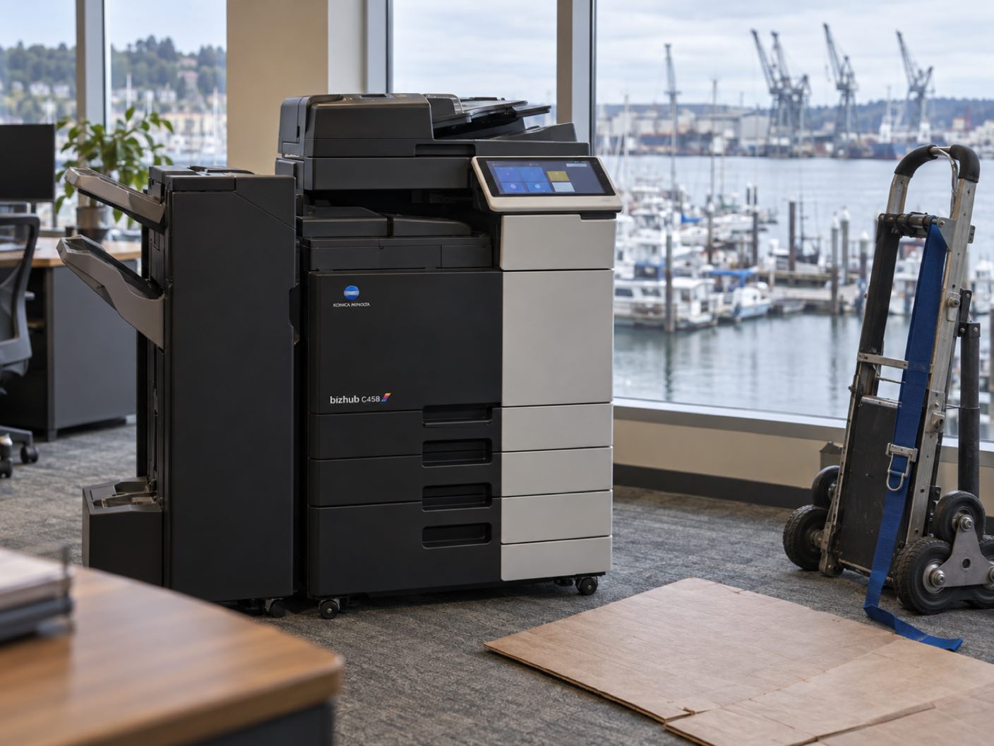 A Konica Minolta bizhub C458 copier newly delivered and standing in position, a delivery trolley and floor protection beside it, in an office over a small waterfront, a marina of moored boats and shipyard cranes across the inlet.