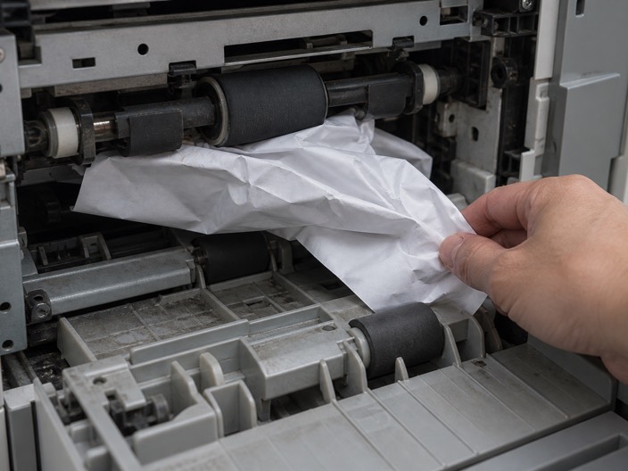 A crumpled sheet of paper being pulled out from between the rollers of an open copier.